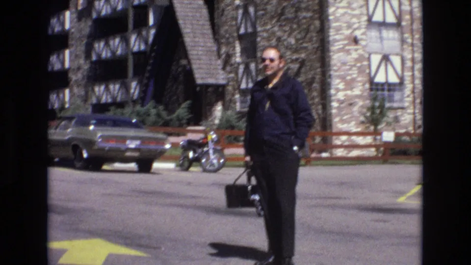 1973 Colorado 8mm frame showing an adult man in dark clothing and sunglasses holding a black briefcase or bag in a parking lot near a classic car, motorcycle, and stone-and-timber buildings.