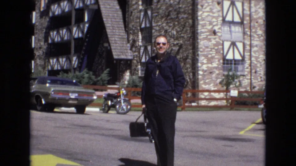 1973 Colorado 8mm frame showing an adult man in sunglasses walking across a parking lot with a black bag, with a classic car, motorcycle, fence, and timber-framed building behind him.