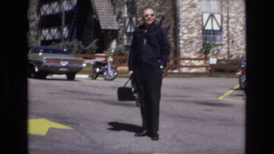 1973 Colorado 8mm frame showing an adult man in dark clothing and sunglasses standing with a black bag in a parking lot, with a classic car, motorcycle, yellow pavement arrow, and stone-and-timber buildings visible.
