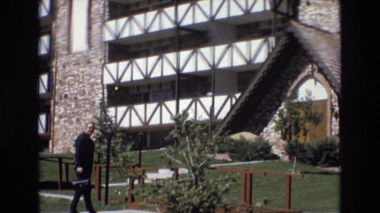 Man in dark clothing walks across a lawn beside a Tudor-style building exterior.