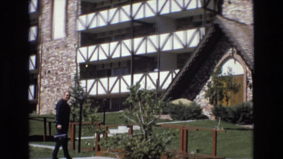 In Colorado home-movie footage from 1973, a man in dark clothing walks left to right across a manicured lawn in front of a multi-story stone and white timber-framed building.