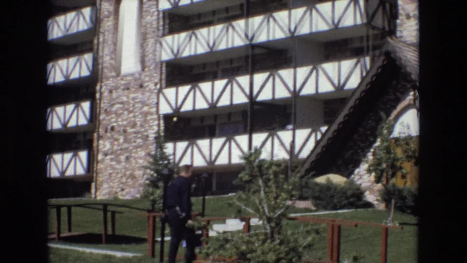 The man continues across the landscaped grounds toward the Tudor-style building, with young trees, shrubs, wooden railings, stonework, and white cross-timbering visible behind him.