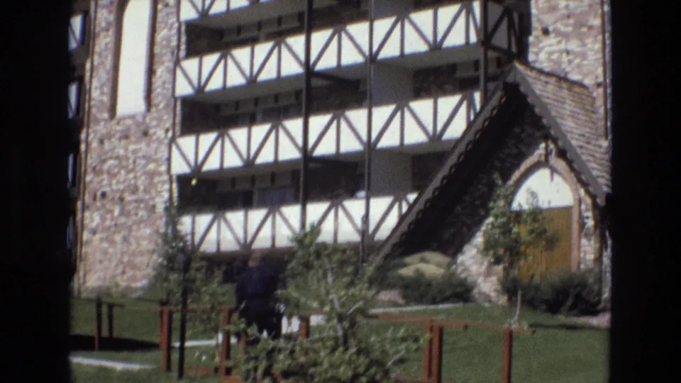 In the lower frame, the man appears partly lowered or obscured by shrubs while crossing the grounds in front of the stone and timber-framed building exterior.