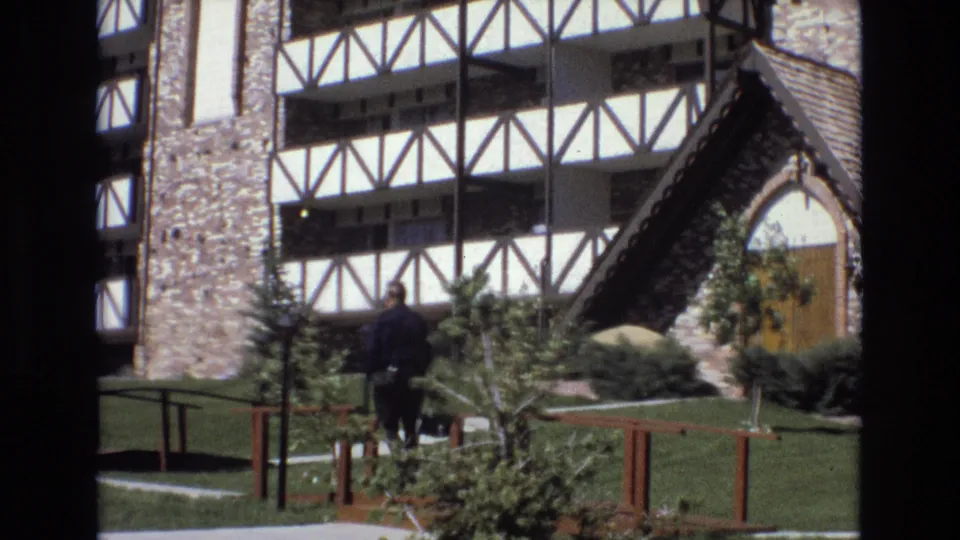 The man is seen from behind as he walks farther across the Colorado landscaped grounds toward the distinctive stone and white timber-framed building and arched roof structure.