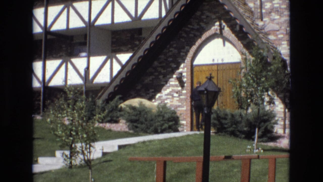 1973 Colorado home-movie frame showing a brick and white half-timbered building with a steep gabled entry, arched wooden door, lawn, shrubs, and a black lamppost.