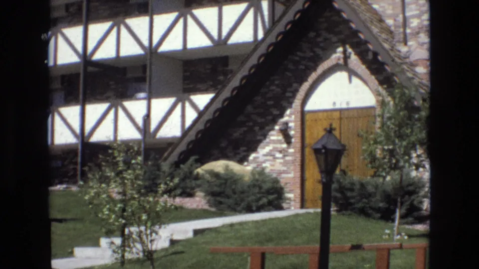 1973 Colorado frame of a chalet-like brick building entrance with half-timbered upper facade, arched wood door, trimmed shrubs, grass, walkway, and a black lamppost in front.