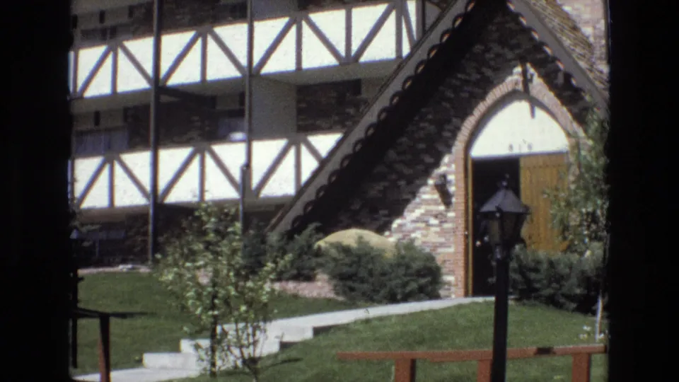 1973 Colorado home-movie view of the steep gabled brick entry, white half-timbering, arched wooden door, concrete steps, small trees, shrubs, lawn, and lamppost.