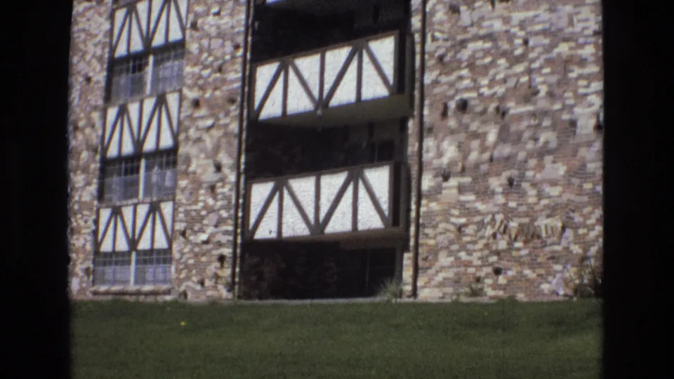 1973 Colorado frame showing the side facade of a brick and half-timbered building with rectangular windows, dark recessed openings, lawn, and textured stone or brickwork.