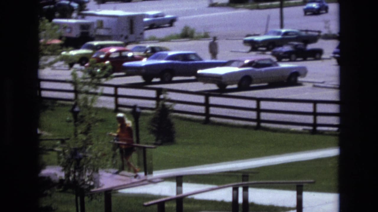 In 1973 Colorado home-movie footage, a woman in a yellow top walks along a landscaped path while classic cars sit in a parking lot beyond a wooden fence.
