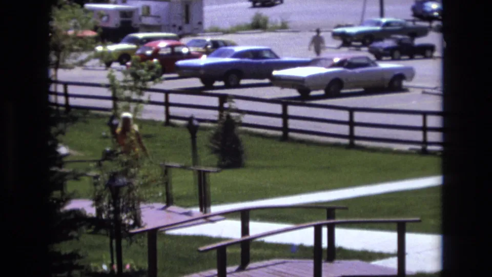 A woman continues along the path near low railings, with a blue car, a white convertible, and another pedestrian visible across the parking lot.