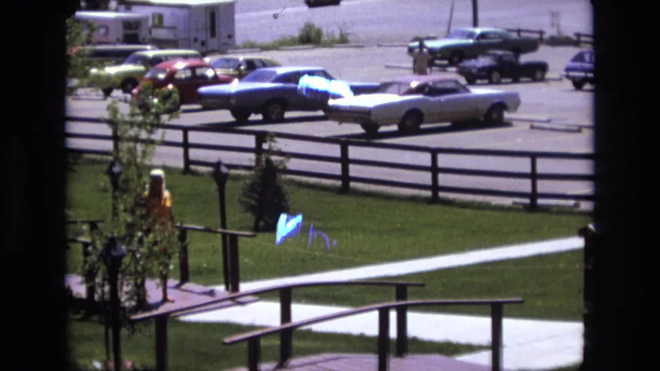The woman is partly obscured by a young tree while walking near the lawn and railing, with several classic cars parked in the bright background.