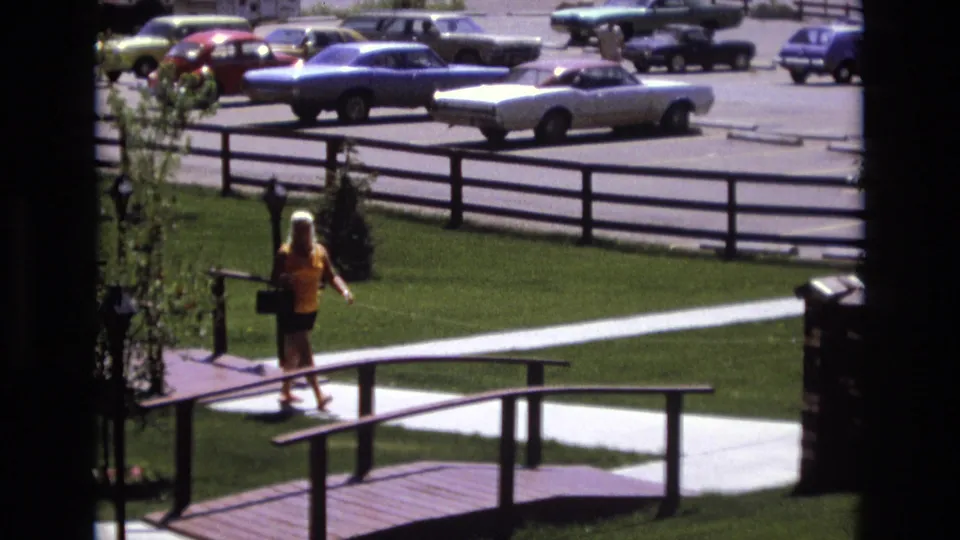 The woman walks farther along the curved path, seen from above through a window frame, with railings, lawn, and a lot of parked 1970s-era cars behind her.