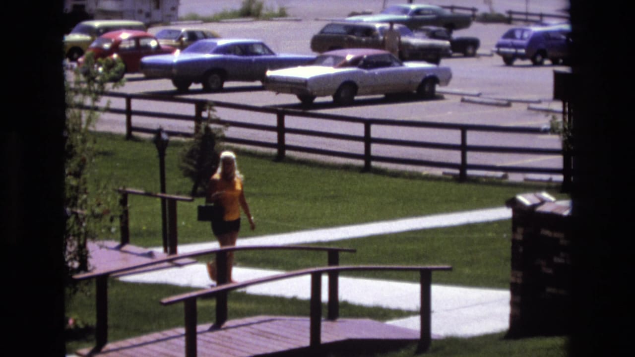 In Colorado 1973 home-movie footage, a blonde woman in a yellow top and dark shorts walks on a paved park path toward a small wooden bridge, with early-1970s cars visible beyond a fence.