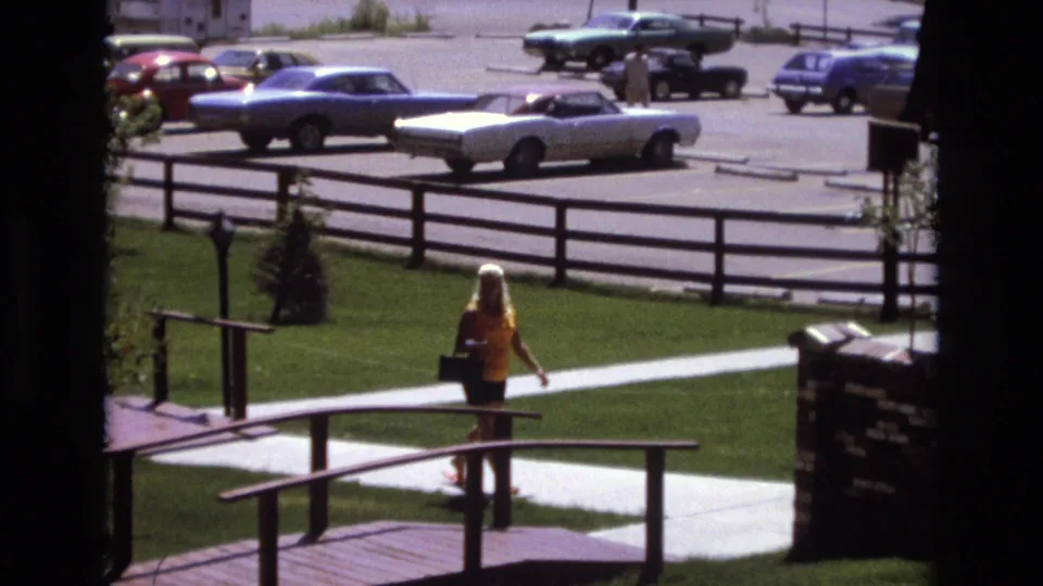 In the 1973 Colorado park scene, the woman continues along the paved path beside wooden bridge railings while a white convertible, blue car, and other parked vehicles sit beyond the fence.