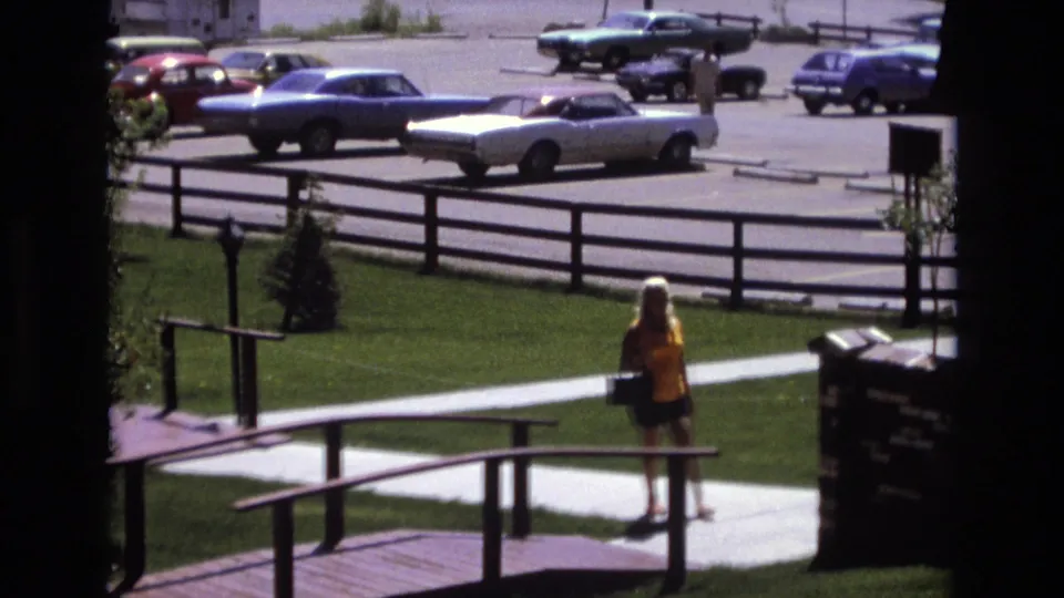 The woman appears near the bridge approach in the Colorado 1973 home-movie frame, with green grass, a paved walkway, wooden rails, and multiple early-1970s cars in the background.