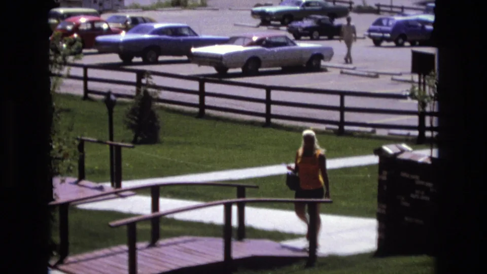 At the end of the short Colorado 1973 park shot, the woman reaches the small bridge area as railings cross the foreground and period cars remain visible across the fence.