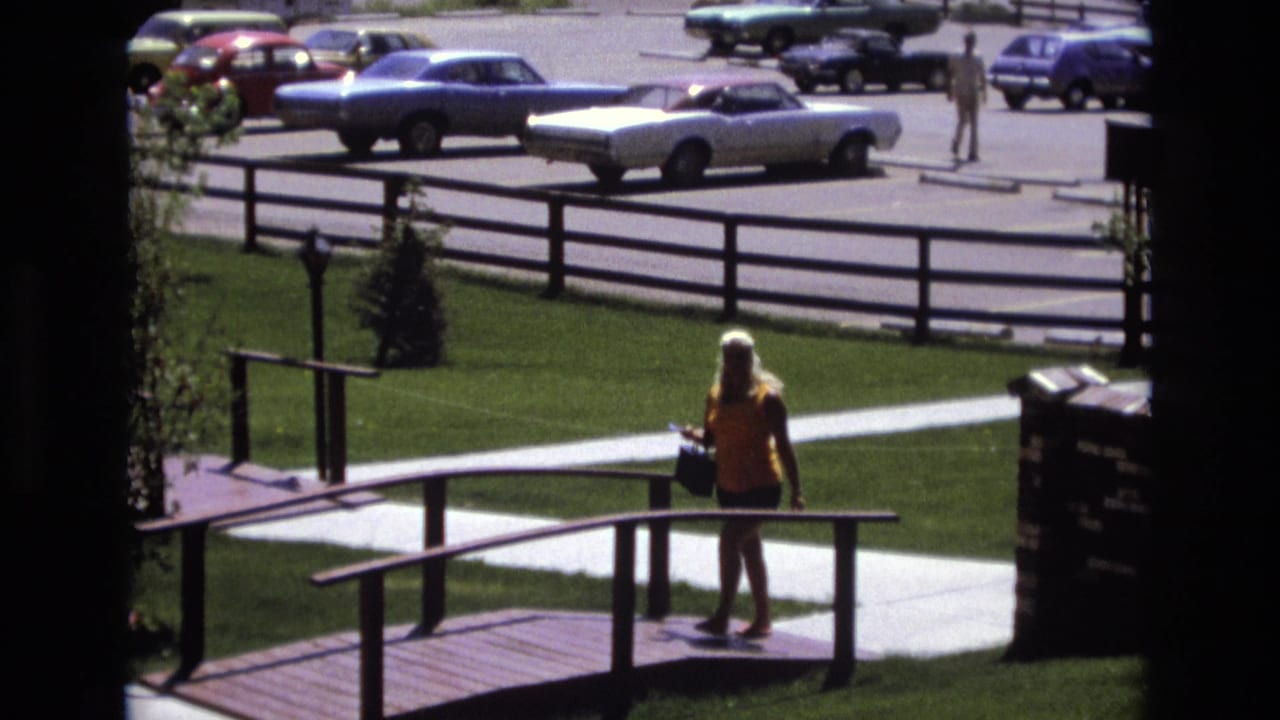 In Colorado home-movie footage from 1973, a woman in a yellow top and dark shorts crosses a wooden ramp with parked classic cars in the background.