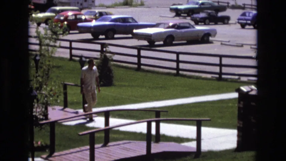 A man in light-colored clothing walks near a landscaped sidewalk and wooden railing, with 1970s parked cars visible across the lot in the background.
