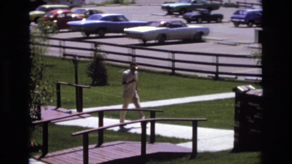 The distant 1973 Colorado 8mm frame shows a man crossing near a wooden bridge or ramp while classic American cars sit in the bright parking lot beyond.