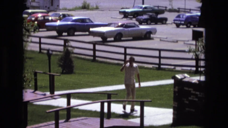 A man in light clothing walks toward the camera from a sidewalk near a wooden ramp, with a fenced parking lot and 1970s cars behind him.