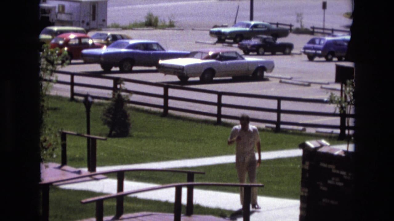In 1973 Colorado home-movie footage, a man in a white shirt and light pants approaches a small wooden bridge or walkway with lawns, fencing, and parked cars behind him.