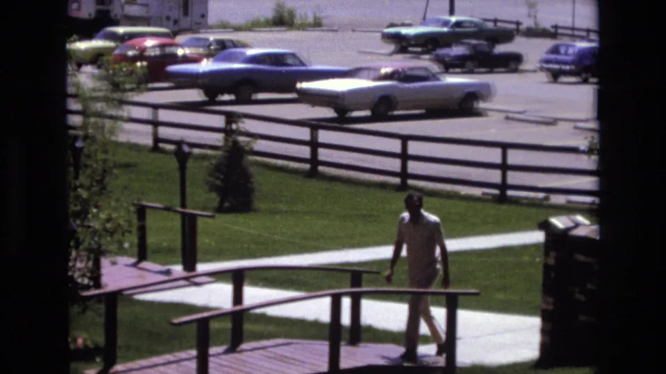 The man continues across the small bridge in a sunlit Colorado suburban setting, with period cars, a sidewalk, lawn, and rail fencing visible behind him.