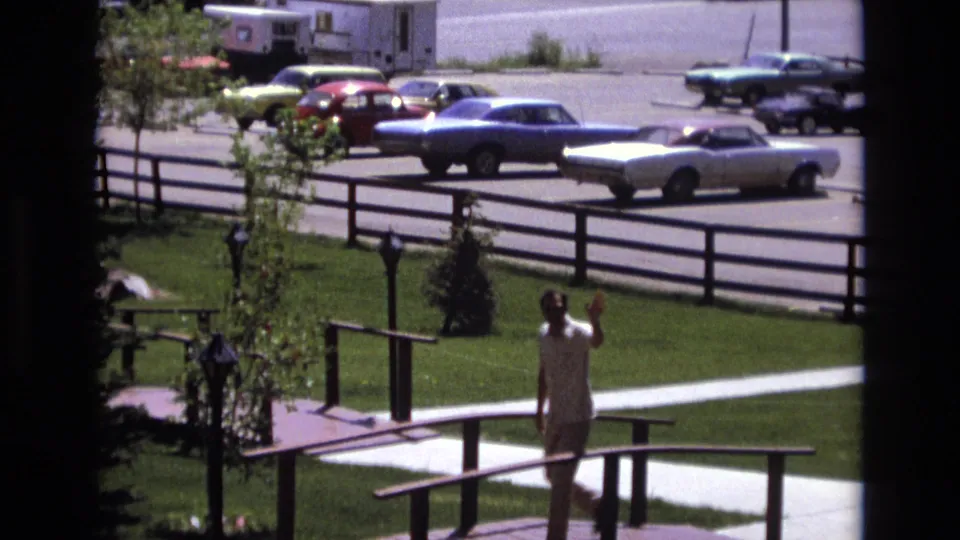 The man raises a hand in a friendly wave while standing or walking on the small bridge, framed by a lawn, young trees, rail fencing, and 1970s parked cars.