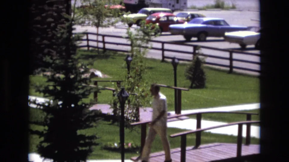 Near the end of the clip, the man moves away along the walkway beside the small bridge, with the Colorado suburban lawn, fence line, shrubs, and parked cars still visible.