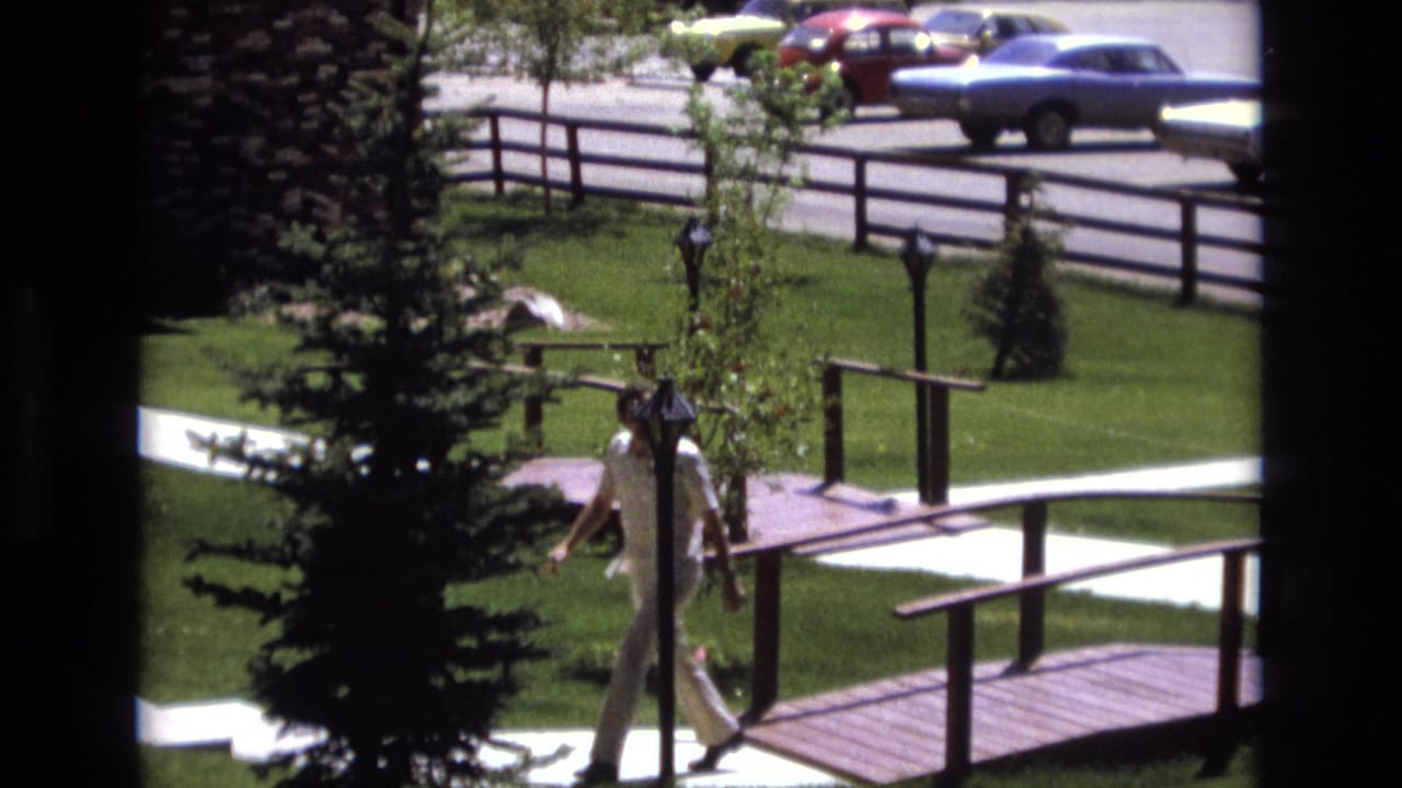 A man in a white shirt and light pants walks through a sunlit Colorado park in 1973, with grass, trees, a wooden bridge, a fence, and older cars visible beyond the grounds.