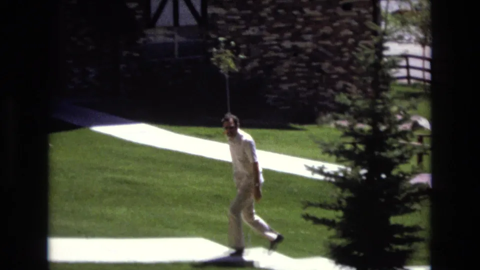 A man wearing sunglasses and light clothing walks across a bright white path in the green Colorado park, with trees and a stone-faced building in the background.