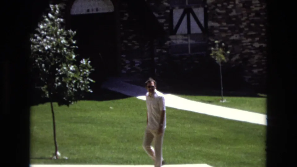 The man appears closer to camera in the 1973 Colorado park, wearing sunglasses and light clothing near a white concrete path, green lawn, young tree, and stone-faced building.
