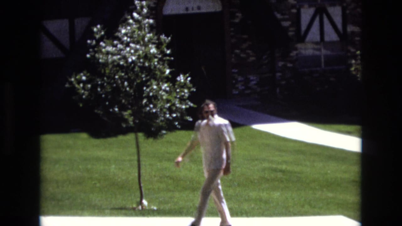 In 1973 Colorado home-movie footage, a man in aviator sunglasses, a white short-sleeved shirt, and beige pants walks across a manicured lawn near a stone-and-timber house facade.