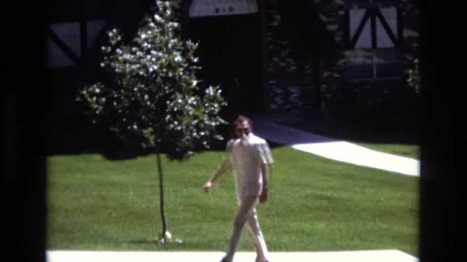 In 1973 Colorado home-movie footage, a man in aviator sunglasses, a white short-sleeved shirt, and beige pants walks across a manicured lawn near a stone-and-timber house facade.