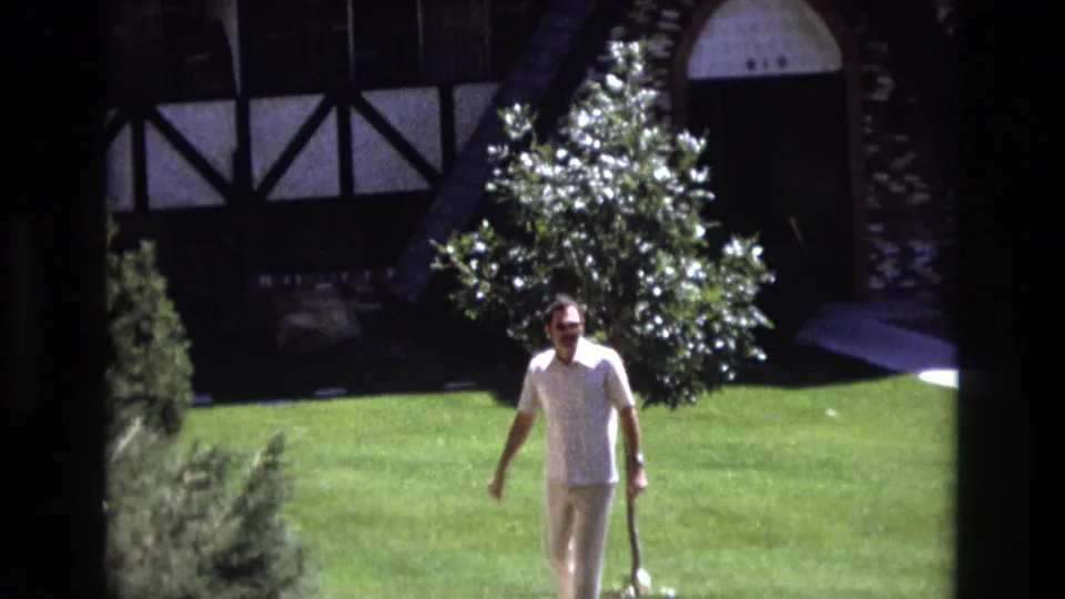 The man continues across the sunlit Colorado lawn, with shrubs, a small tree, a paved walkway, and the Tudor-style house exterior visible behind him.