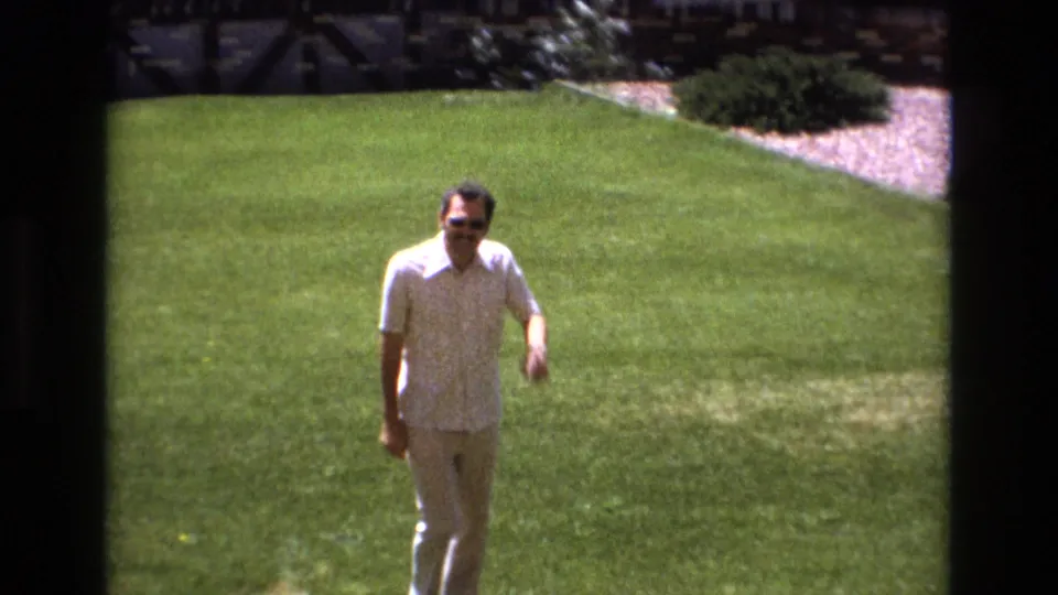 A closer frame shows the man approaching the camera on the manicured lawn, wearing sunglasses and light 1970s casual clothing.