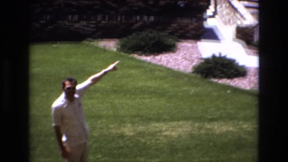 The man pauses on the Colorado lawn and points toward something outside the frame, with shrubs, walkway, and part of the house grounds in the background.