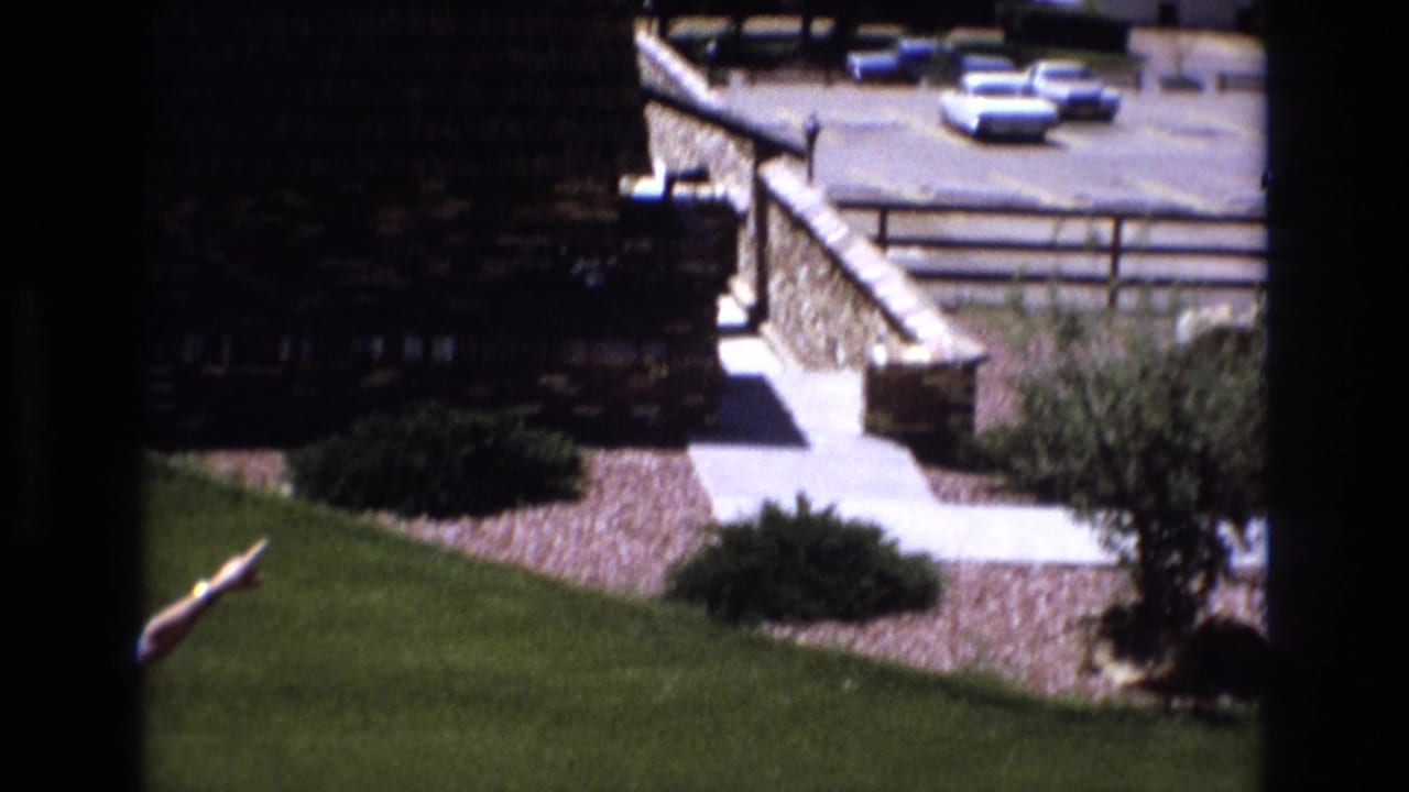 Colorado 1973 home-movie frame showing a stone retaining wall, paved walkway, green lawn, shrubs, and several parked cars at a mountain resort area.