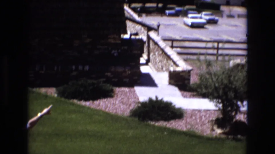 Colorado 1973 home-movie frame showing a stone retaining wall, paved walkway, green lawn, shrubs, and several parked cars at a mountain resort area.