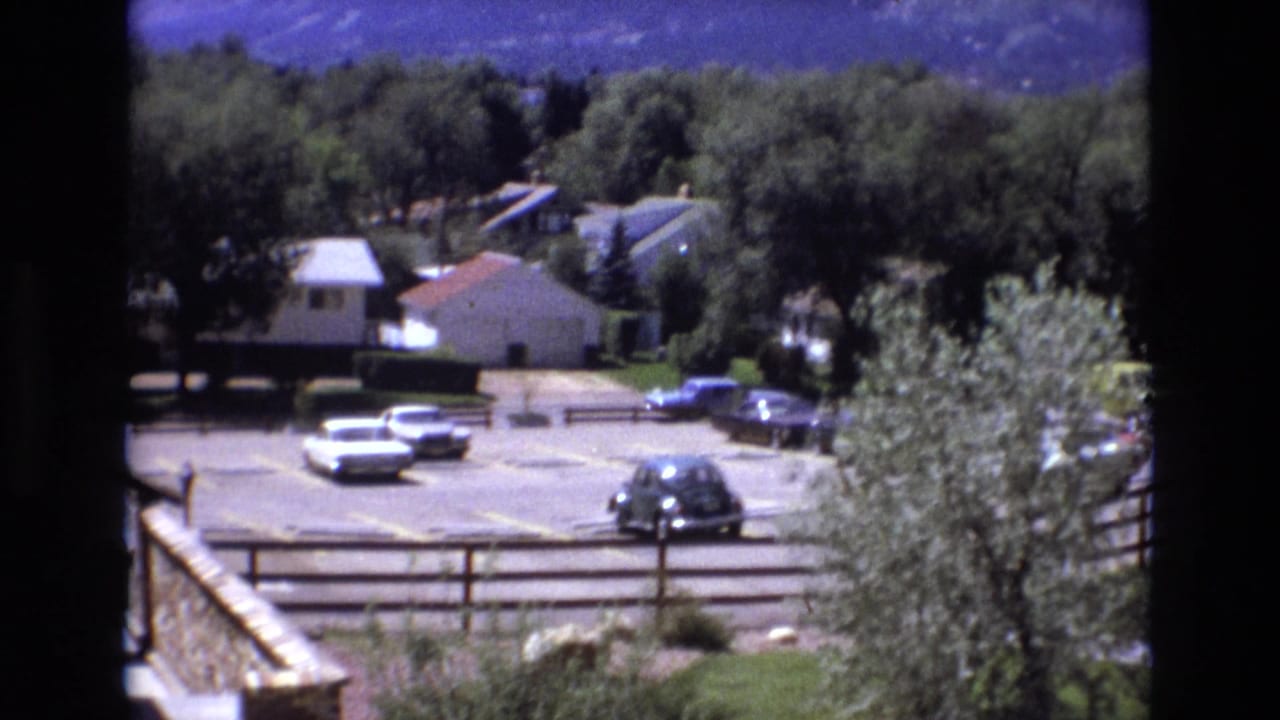 Colorado 1973 resort-area view with a parking lot, period cars, small buildings, trees, fencing, and a mountain ridge beyond.