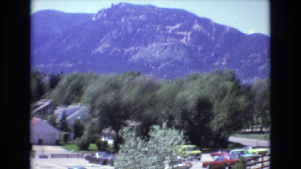 Colorado 1973 travel frame looking across trees, rooftops, parked cars, and fencing toward a large mountain ridge under blue sky.