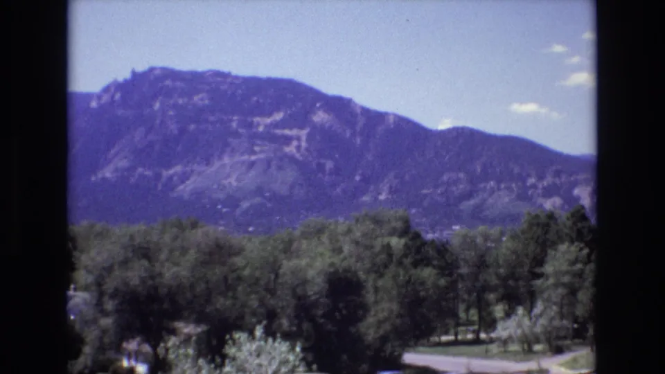 Colorado 1973 scenic home-movie frame with a broad mountain ridge, dense trees, pale blue sky, and a small road or parking area below.