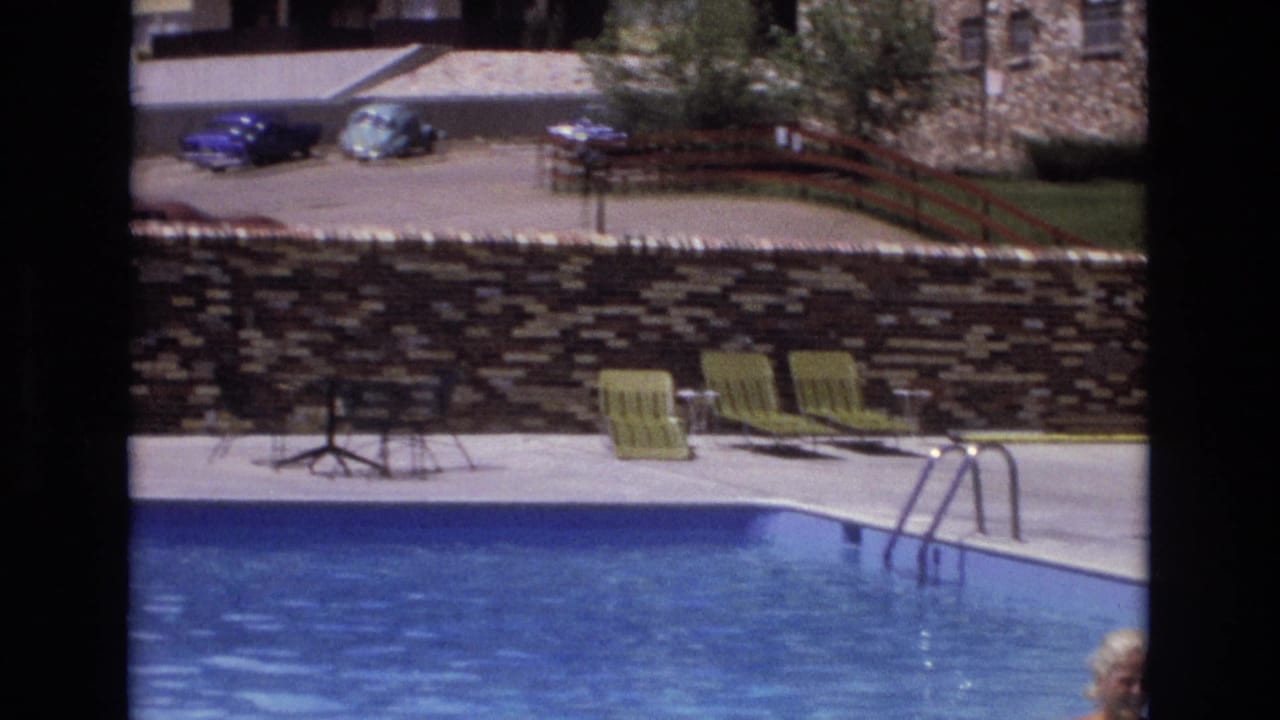 Poolside patio with blue water, green lounge chairs, stone wall, parked cars, and a partial person at the frame edge.