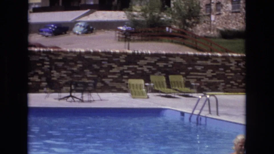 1973 Colorado home-movie frame showing a resort pool, green lounge chairs, a stone retaining wall, parked cars beyond it, and a partially visible person near the right edge.