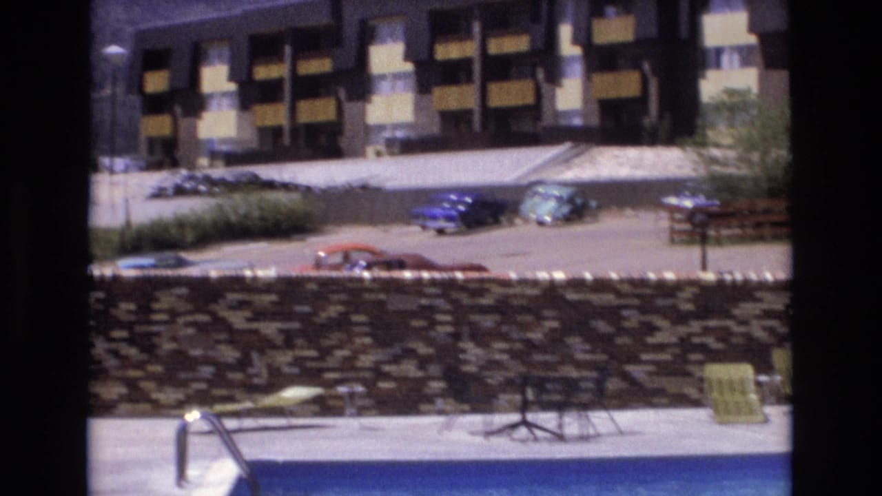 Pool edge, metal ladder, stone wall, parked cars, bench, and multi-story building in bright daylight.