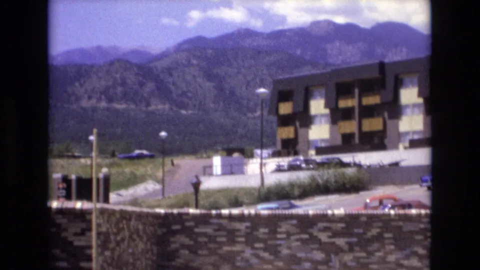 1973 Colorado home-movie view of forested mountains rising behind a multi-story resort or motel building, with parked cars and lamp posts in the foreground.
