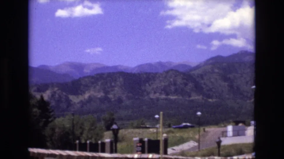 1973 Colorado frame looking past a resort parking area toward layered forested mountains under a blue sky with scattered white clouds.