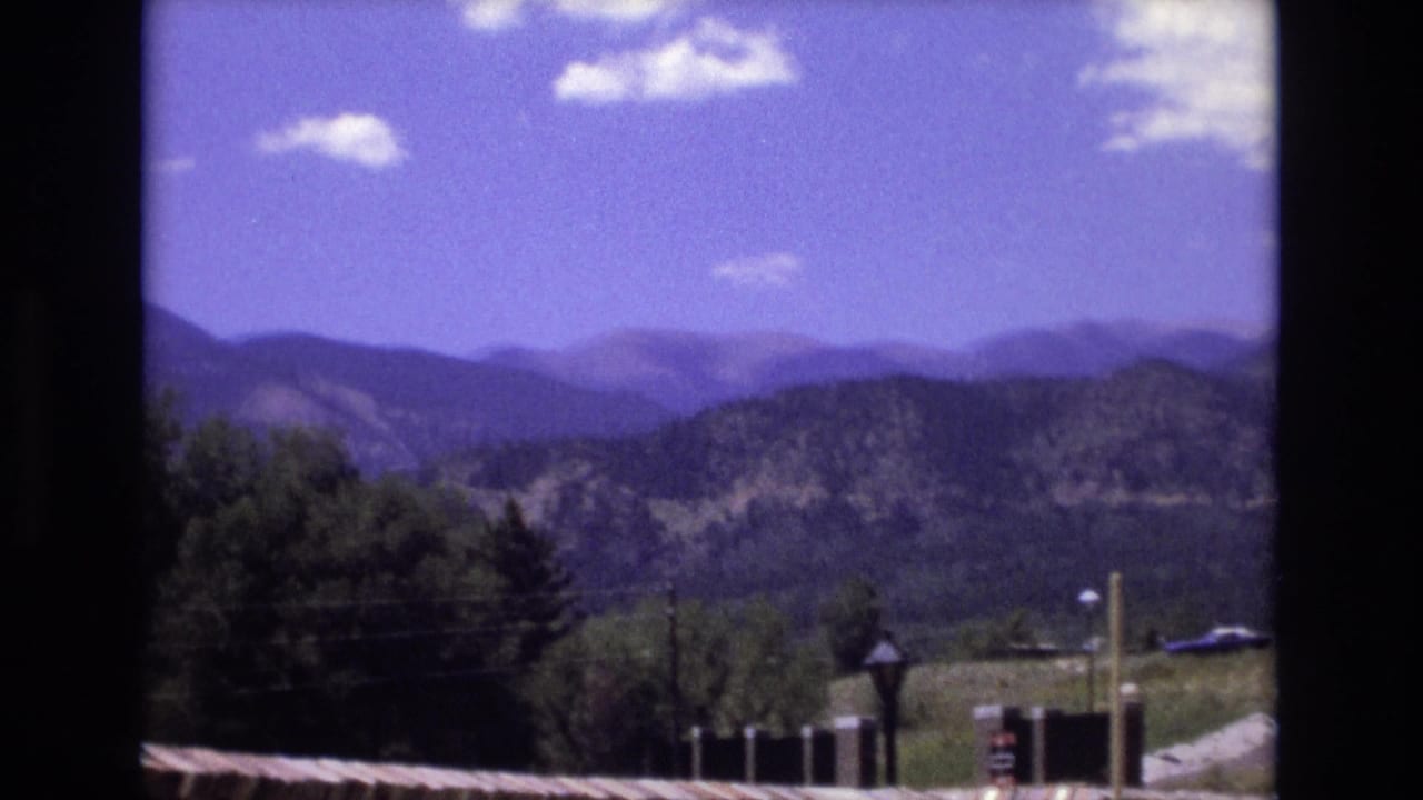 1973 Colorado 8mm frame showing distant mountain ridges under a bright blue sky, with trees, a roof edge, and dark framing at the sides.