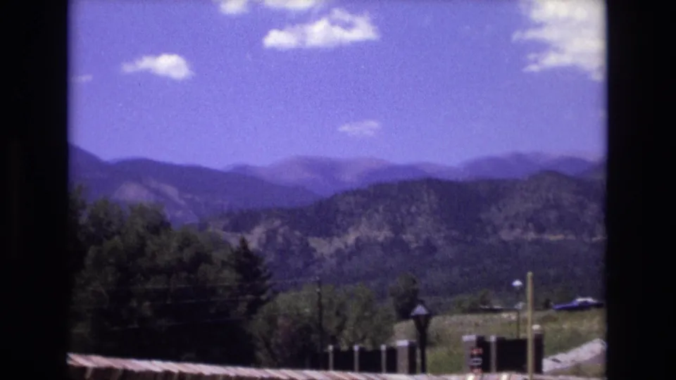 1973 Colorado 8mm frame showing distant mountain ridges under a bright blue sky, with trees, a roof edge, and dark framing at the sides.