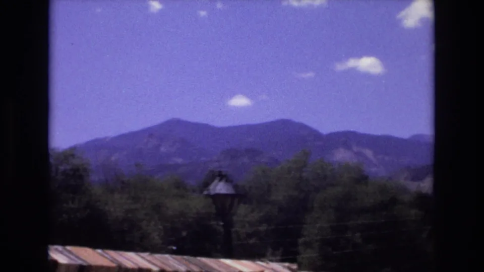 1973 Colorado 8mm frame of a mountain range and treetops seen through a dark border, with a small lamp post and roof edge in the foreground.