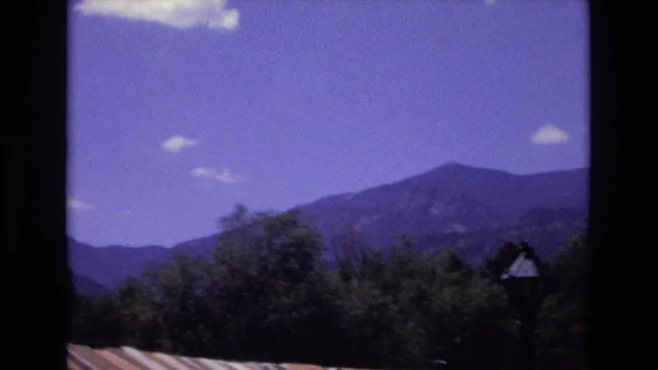 1973 Colorado 8mm frame showing a broad mountain slope behind foreground trees, with a roof edge and dark framing visible along the lower and side edges.